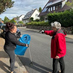 Zwei Frauen sammeln Müll in einer Wohngegend, während eine einen blauen Sack hält und die andere mit einer Zange Müll aufhebt.