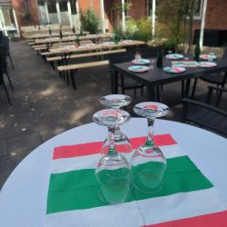 Drei umgedrehte Weingläser stehen auf einer italienischen Flagge, umgeben von festlich gedeckten Tischen im Freien.