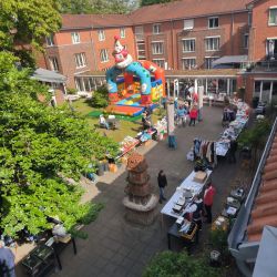 Ein bunter Flohmarkt mit Hüpfburg belebt einen sonnigen Innenhof zwischen roten Backsteinbauten, voller Stände und Besucher.