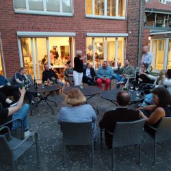 Eine Gruppe von Menschen sitzt abends im Kreis im Freien und unterhält sich lebhaft bei Getränken vor einem beleuchteten Haus.