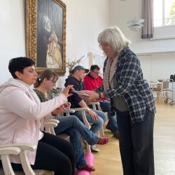 Eine Frau verteilt Dokumente an eine sitzende Gruppe in einem Raum mit großem Gemälde an der Wand.
