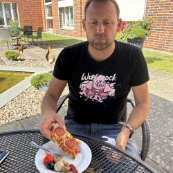 Ein Mann genießt draußen an einem Tisch ein Stück Pizza und Antipasti vor einem roten Backsteingebäude.