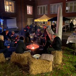 Menschen sitzen abends gemütlich auf Strohballen um ein Lagerfeuer und genießen ein geselliges Beisammensein im Freien.