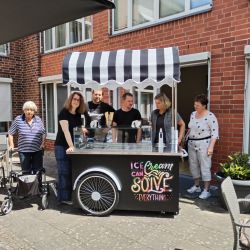 Eine Gruppe von Menschen versammelt sich fröhlich um einen Eiswagen mit gestreiftem Dach vor einem roten Backsteingebäude.