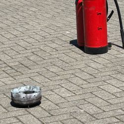 Ein roter Feuerlöscher und ein kleiner Metallbehälter stehen auf einem sonnigen, gepflasterten Boden.