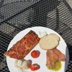 Auf einem weißen Teller im Sonnenschein liegen Pizza, Lachs mit Dill, Brot, Paprika und Pilze auf einem Metalltisch.