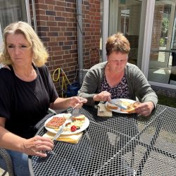 Zwei Frauen genießen bei sonnigem Wetter eine Mahlzeit an einem Metalltisch im Freien.