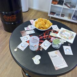 Ein Tisch aus einem Coca-Cola-Fass ist mit Chips, Karten und Schreibutensilien dekoriert und lädt zum Verweilen ein.