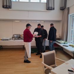 Eine Gruppe von Männern steht in einem Raum mit Buffet, während einer einen Teller von einem Mann in roter Kleidung überreicht bekommt.