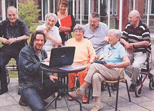 Ein Mann erklärt einer Gruppe älterer Menschen im Freien etwas am Laptop, während sie aufmerksam bei der Fortbildung zuhören.