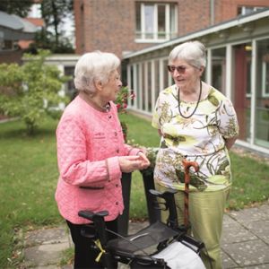 Zwei ältere Frauen unterhalten sich fröhlich im Garten; eine stützt sich auf einen Rollator, die andere auf einen Spazierstock.