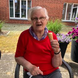 Eine ältere Frau genießt ein Eis in der Sonne, umgeben von blühenden Blumen vor einer Backsteinfassade.