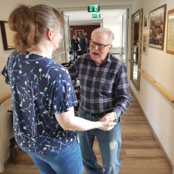 Ein älterer Mann und eine Frau halten sich lächelnd an den Händen in einem hell erleuchteten Flur voller Bilder.