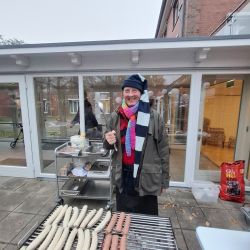 Eine Person steht lächelnd am Grill mit Würstchen, eingehüllt in einen Schal und Mütze an einem kühlen Tag.