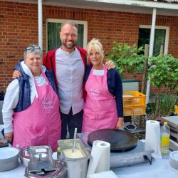 Drei Personen stehen fröhlich vor einem Tisch mit Waffeleisen, während zwei Frauen in rosa Schürzen lächeln.