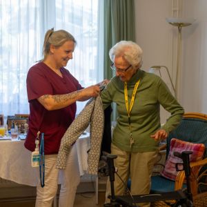Eine Pflegekraft hilft einer älteren Dame beim Anziehen eines Oberteils in einem gemütlichen Wohnraum. | © Name
