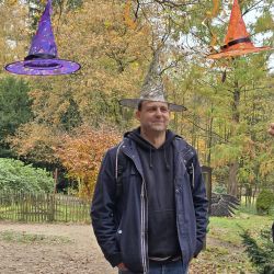 Ein Mann steht lächelnd im Freien und trägt einen glitzernden Hut unter bunt dekorierten Hexenhüten an einem Baum.