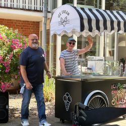Zwei Männer stehen am Eiscafé, während der Betreiber freundlich winkt und die Sonne scheint. | © Name