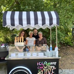 Vier Personen stehen hinter einem dekorativen Eiswagen im Freien und bieten verschiedene Eissorten an, umgeben von Bäumen.