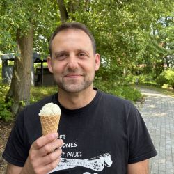 Ein Mann steht lächelnd im Grünen, hält ein Eis und trägt stolz ein schwarzes "St. Pauli"-T-Shirt.