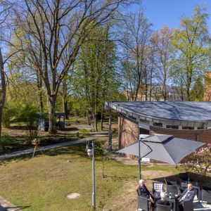Zwei Männer entspannen im Freien auf einer Terrasse, umgeben von Bäumen und modernem Architekturdesign. | © Name