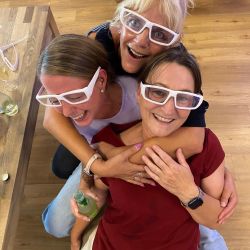 Drei lachende Frauen mit weißen Brillen umarmen sich fröhlich auf einem Holzboden und genießen den Moment der Freundschaft.