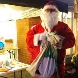 Ein Weihnachtsmann in rotem Anzug hält einen vollen Geschenkesack vor einem festlich beleuchteten Stand auf einem Weihnachtsmarkt.