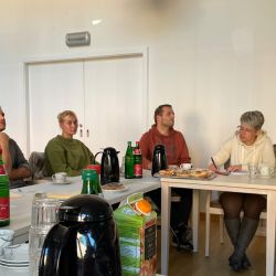 Menschen sitzen an einem Tisch bei einem Meeting, umgeben von Getränken und Snacks, während sie aufmerksam zuhören.