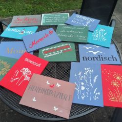 Bunte Schilder mit norddeutschen Sprüchen und Motiven liegen auf einem Tisch im Freien. | © Name