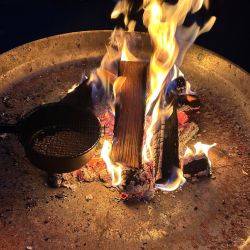 Ein lebhaftes Lagerfeuer brennt in einer Feuerschale mit glühenden Holzscheiten und einem Grillrost.