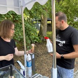 Unter einem Sonnenschirm im Park reicht eine Frau einem Mann ein Eis mit Sahne.