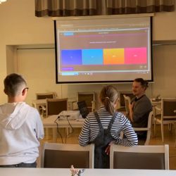 Ein Mann hält am 3. April eine Präsentation vor zwei Kindern in einem Raum mit Projektor und dekoriertem Tisch.