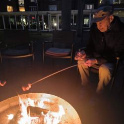 Ein älterer Mann röstet Marshmallows über einem Lagerfeuer im Innenhof und hält dabei ein Getränk in der Hand.