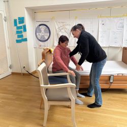 Eine Frau hilft einer anderen beim Aufstehen von einem Stuhl, während Lehrmaterialien an der Wand hängen.
