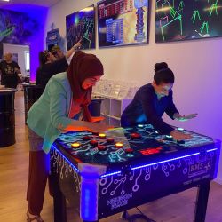 Zwei Frauen spielen konzentriert am leuchtenden Neuron-Race-Tisch, während bunte Bildschirme im Hintergrund strahlen.