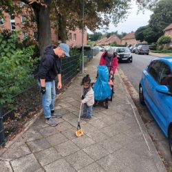 Ein Kind sammelt mit einer Greifzange Müll auf dem Gehweg, während ein Erwachsener und ein Rollator vorbeigehen.