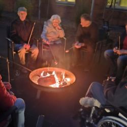Eine Gruppe von Menschen sitzt gemütlich im Kreis und röstet Marshmallows an einem Lagerfeuer bei Abenddämmerung.