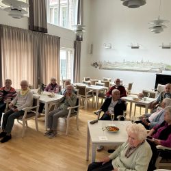 Ältere Menschen sitzen in einem hellen Raum mit Tischen und genießen Snacks und Getränke. | © Name
