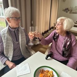 Zwei Frauen stoßen mit Sektgläsern an, während sie an einem Tisch mit Snacks sitzen.  | © Name