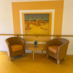 Zwei braune Sessel und ein runder Tisch mit Blumen stehen vor einem gelben Wandbild mit Landschaftsmotiv.