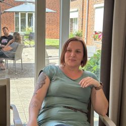 Eine Frau sitzt drinnen entspannt vor einem Stück Pizza, während draußen Menschen unter einem Sonnenschirm plaudern.