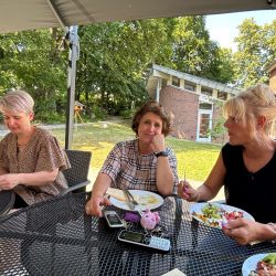 Drei Frauen genießen ein gemütliches Mittagessen unter einem Pavillon im Garten an einem sonnigen Sommertag.