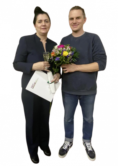 Eine Frau hält ein Sektglas und Blumen, während sie mit einem Mann posiert, der ebenfalls einen Blumenstrauß hält.