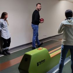 Auf der Kegelbahn steht eine Person mit roter Kugel bereit, während zwei andere interessiert zuschauen.
