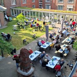Menschen genießen ein Sommerfest im Innenhof eines Seniorenheims mit Musik und Essen unter strahlend blauem Himmel.