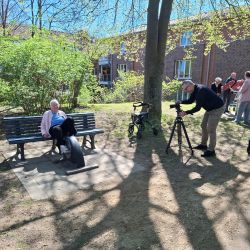 Eine ältere Dame sitzt auf einer Bank im Park, während ein Mann mit einer Kamera filmt. | © Name
