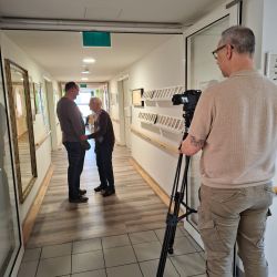 Zwei Personen sprechen in einem Flur, während ein Kameramann sie mit einem Stativ filmt. | © Name