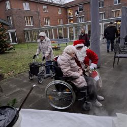 Ein Weihnachtsmann besucht eine ältere Dame im Rollstuhl vor einem Backsteingebäude, umgeben von weiteren Senioren.