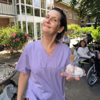 Eine Frau in violetter Arbeitskleidung lächelt draußen und hält eine Schale Eis mit einem Herzkeks.