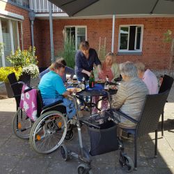 Seniorinnen genießen bei sonnigem Wetter ein gemeinsames Essen im Innenhof und sitzen gemütlich an einem Tisch zusammen.